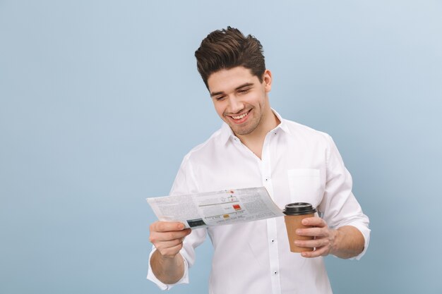 Portrait d'un joyeux beau jeune homme debout isolé sur bleu, tenant une tasse de café à emporter, lisant le journal