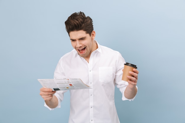 Portrait d'un joyeux beau jeune homme debout isolé sur bleu, tenant une tasse de café à emporter, lisant le journal