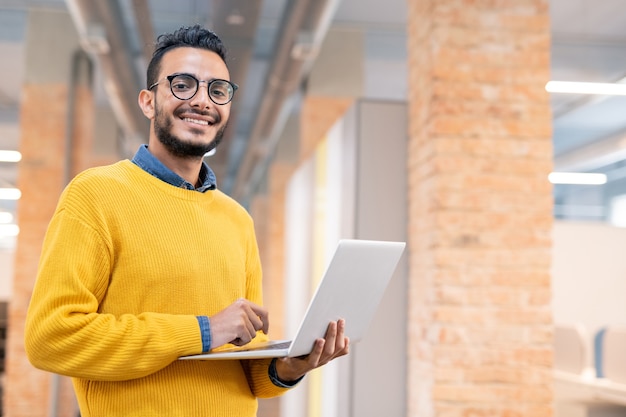 Portrait de joyeux bel homme de bureau métisse avec barbe debout dans l'espace loft et à l'aide d'un ordinateur portable