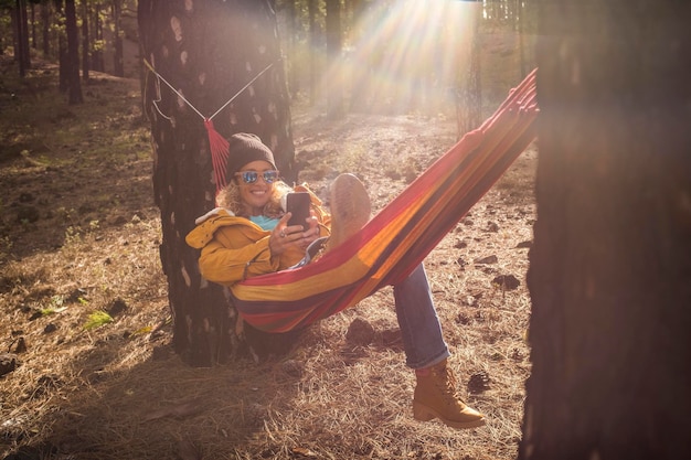 Portrait joyeux d'une belle femme profiter de la détente à la montagne allongée sur un hamac sur la nature forêt bois parc extérieur et souriant à la connexion d'itinérance du téléphone cellulaire et au travail intelligent