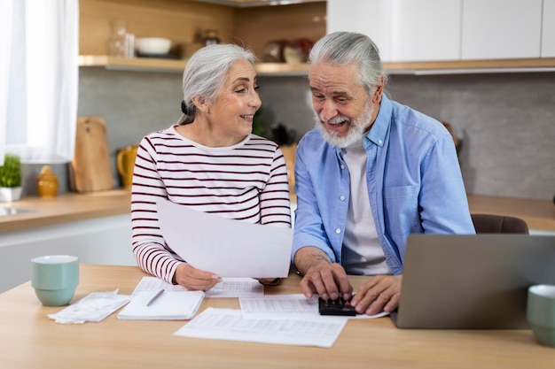 Portrait de joyeux conjoints âgés assis dans la cuisine et vérifiant les papiers financiers