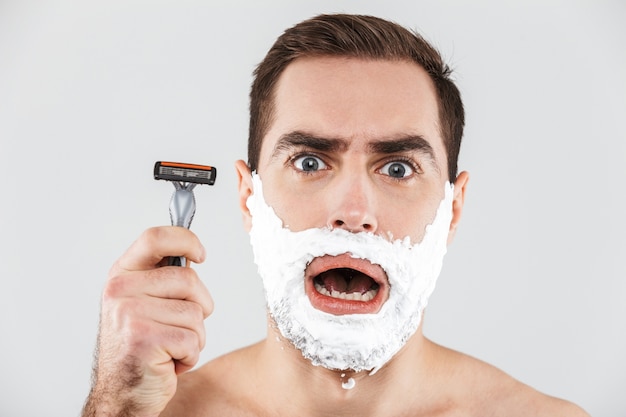 Photo portrait d'un joyeux homme barbu torse nu debout isolé sur blanc, le visage couvert de mousse à raser, tenant un rasoir