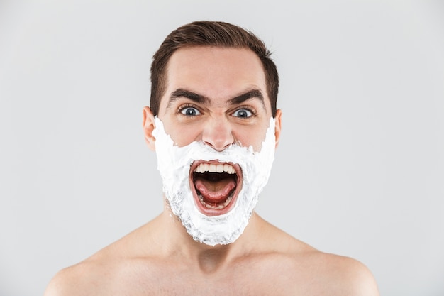 Portrait d'un joyeux homme barbu torse nu debout isolé sur blanc, le visage recouvert de mousse à raser