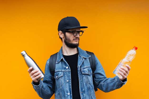 Portrait de joyeux jeune hipster, tenant une bouteille en acier thermo eco pour eau et bouteille en plastique, sur un mur jaune ou orange. Dites non au plastique.