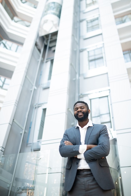 Portrait de joyeux jeune homme d'affaires afro-américain en costume debout avec les bras croisés dans le hall du bureau