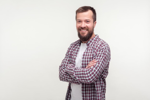 Portrait de joyeux jeune homme barbu de bonne humeur en chemise à carreaux décontractée croisant les bras et souriant sincèrement à la caméra, l'air satisfait, satisfait de la vie. tourné en studio isolé sur fond blanc