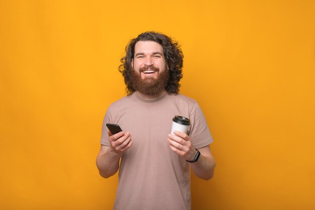 Portrait de joyeux jeune homme barbu souriant tenant smartphone et café à emporter