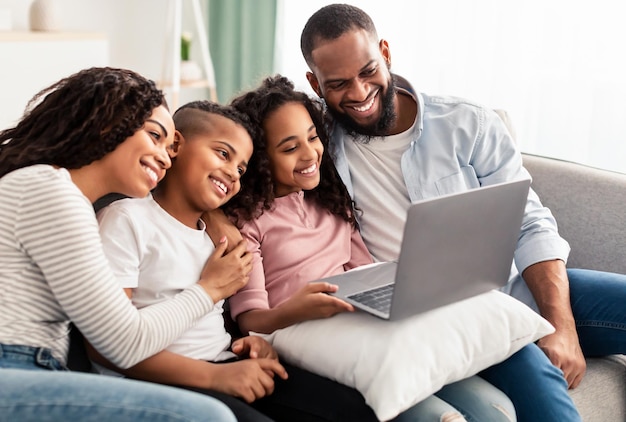 Portrait de joyeux parents afro-américains et de leurs enfants heureux utilisant un ordinateur portable sur un oreiller, regardant un film ou ayant une vidéoconférence en ligne, assis sur le canapé dans le salon à la maison ensemble