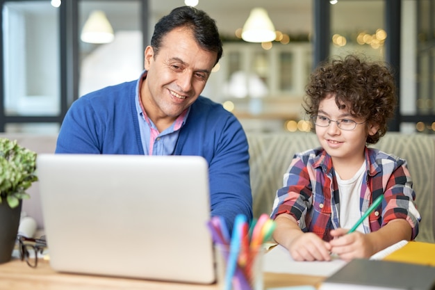 Portrait de joyeux père hispanique d'âge moyen passant du temps avec son fils. Petit garçon assis au bureau avec son père et fait ses devoirs, à l'aide d'un ordinateur portable. Étude en ligne, concept de famille