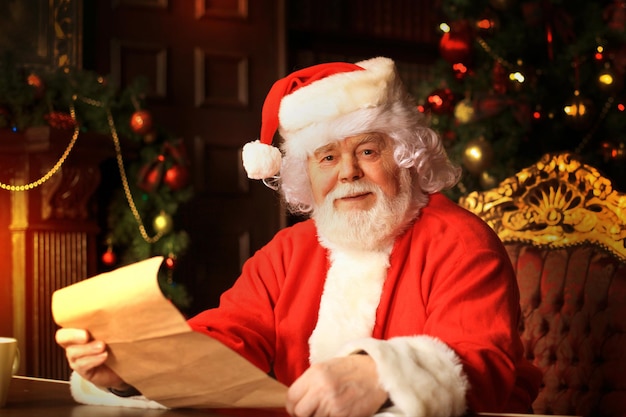 Portrait d'un joyeux père Noël assis dans sa chambre à la maison près de l'arbre de Noël et lisant une lettre de Noël ou une liste de souhaits.