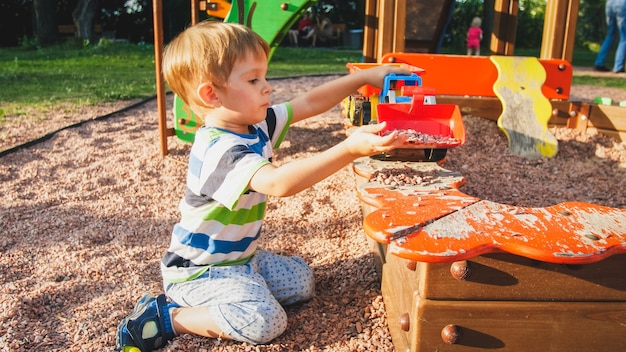 Portrait de joyeux petit garçon souriant versant du sable dans un camion jouet avec remorque. Enfants jouant et ayant sur aire de jeux au parc