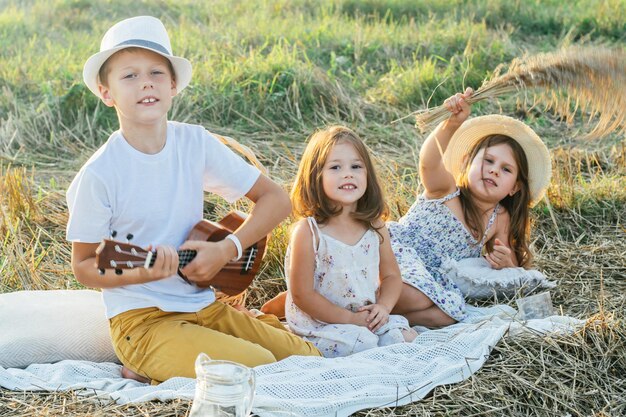 Portrait de joyeux trois enfants assis sur une couverture dans un champ jouant au ukulélé en train de pique-niquer avec des boissons et des plats savoureux