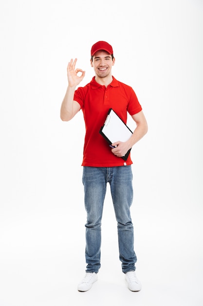 Portrait de livreur caucasien en t-shirt rouge et casquette souriant et montrant le symbole bien avec les doigts tout en tenant le presse-papiers, isolé sur l'espace blanc