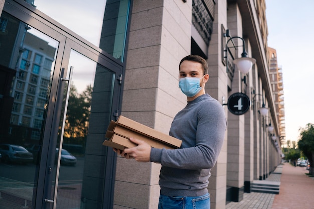 Portrait de livreur en masque qui sonne à la porte pour les boîtes de livraison avec pizza en regardant la caméra