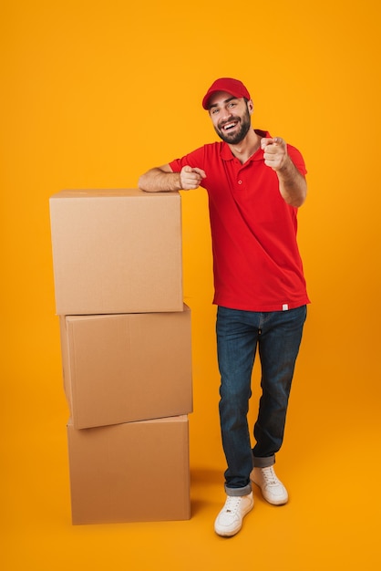 Portrait d'un livreur optimiste en uniforme rouge pointant le doigt vers la caméra en se tenant debout avec des boîtes d'emballage isolées sur du jaune