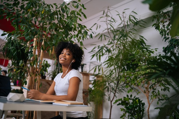 Portrait Lowangle d'une femme afro-américaine assez bouclée écoutant de la musique dans des écouteurs assis à table avec un ordinateur portable et souriant regardant la caméra