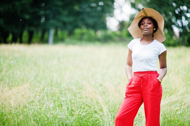 Portrait d'une magnifique femme afro-américaine des années 20 portant un chapeau d'été, un pantalon rouge et un t-shirt blanc posant sur l'herbe verte dans le parc