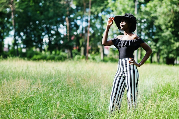 Portrait d'une magnifique femme afro-américaine des années 20 vêtue d'un pantalon à rayures noires et blanches et d'un chapeau d'été posant sur l'herbe verte dans le parc.