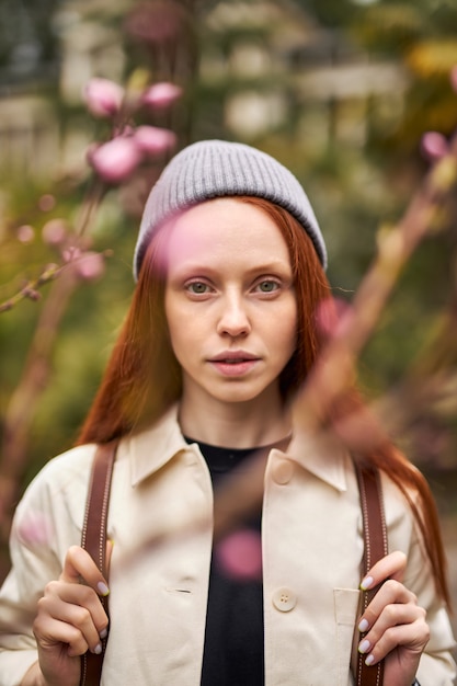 Portrait d'une magnifique femme caucasienne posant au printemps ou en automne à l'extérieur, debout parmi les arbres en fleurs en regardant la caméra avec confiance, une femme aux cheveux roux pose, en chapeau et manteau