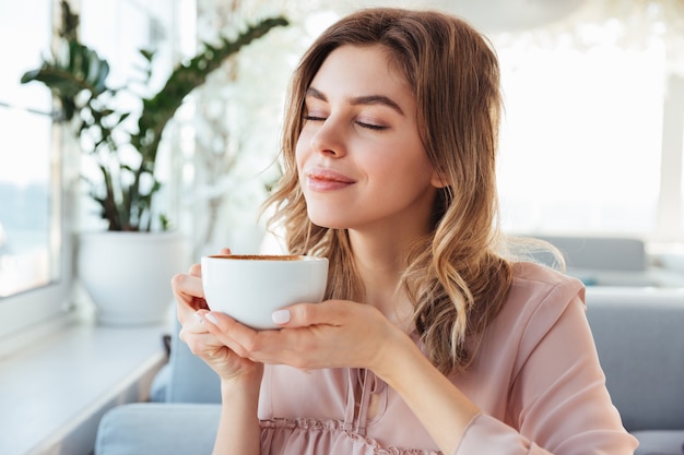 Portrait de magnifique femme souriante sentant et buvant du cappuccino de tasse, tout en se reposant au restaurant le matin