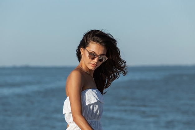 Portrait d'une magnifique fille brune en robe blanche et lunettes de soleil près de la mer par une journée ensoleillée. .