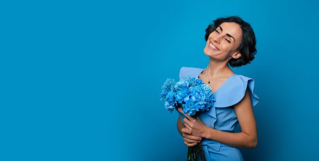 Le portrait d'une magnifique jeune femme heureuse et excitée dans une élégante robe bleue pose avec un bouquet frais de fleurs bleues. Fête des mères. Les vacances des femmes. Printemps. Les droits des femmes