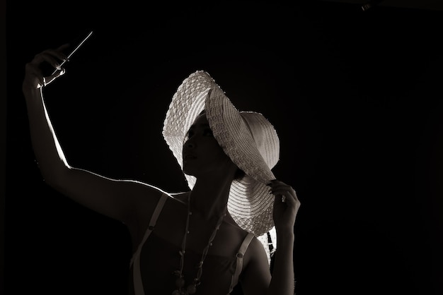 Portrait maillot de bain d'une femme asiatique des années 20 dans l'ombre à faible luminosité avec rétro-éclairage, vue arrière latérale du corps de torsion de la femme prendre une photo de selfie avec un style de chapeau de mode de téléphone. fumée sur fond noir