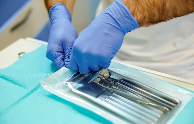 Un portrait des mains d'un dentiste ouvrant un sac stérile avec des outils stériles. Un sac contient un miroir buccal et une pince en coton