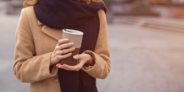 Portrait des mains d'une femme tenant une tasse de café portant un manteau beige et une écharpe noire debout