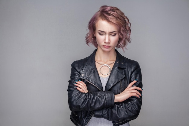 Portrait De Malheureuse Triste Belle Fille Avec Une Coiffure Courte Et Un Maquillage Dans Une Veste En Cuir Noir De Style Décontracté Debout Les Yeux Fermés Avec Les Bras Croisés. Tourné En Studio Intérieur, Isolé Sur Fond Gris.