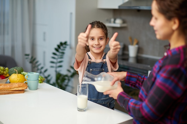 Portrait d'une maman verse son lait de fille mignonne en verre pour le petit déjeuner, petite fille montre les pouces à la caméra.