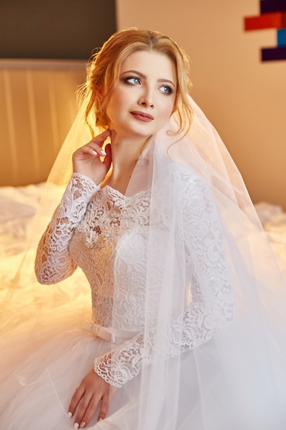 Portrait d'une mariée assise dans une robe de mariée blanche chic sur le lit et se préparant à la cérémonie de mariage. Une femme blonde en robe blanche et un voile sur la tête. La mariée attend son fiancé