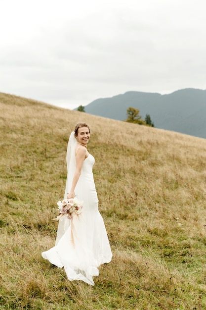 Portrait de mariée Une jeune fille vêtue d'une robe de mariée blanche et avec un bouquet de fleurs et de verdure dans ses mains sur fond de montagnes et de forêts au coucher du soleil