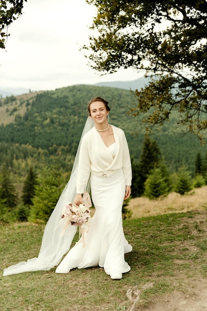 Portrait de mariée Une jeune fille vêtue d'une robe de mariée blanche et avec un bouquet de fleurs et de verdure dans ses mains sur fond de montagnes et de forêts au coucher du soleil