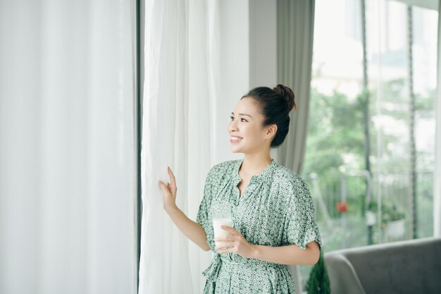 Portrait matinal d'une jeune femme souriante buvant du lait du verre femme debout à la maison près de la fenêtre manger des aliments sains