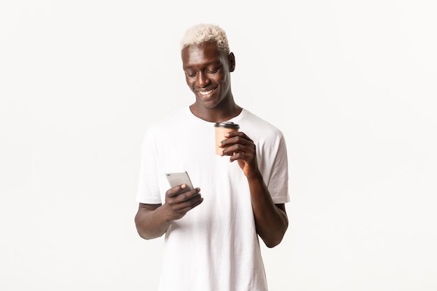 Portrait de mec afro-américain blond moderne élégant, messagerie, à l'aide de téléphone portable et de boire du café à emporter