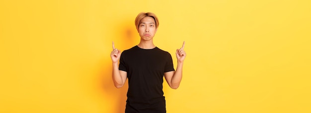 Portrait d'un mec asiatique sombre et boudeur qui a l'air déçu pointant du doigt vers le haut sur fond jaune