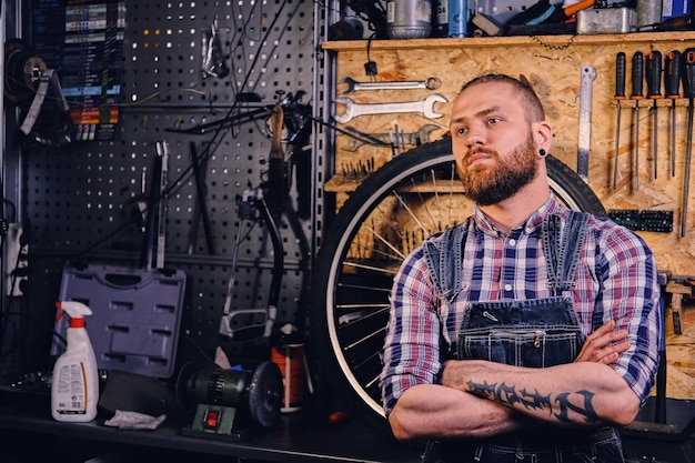 Portrait d'un mécanicien de vélo barbu à tête rouge dans un atelier avec des pièces de vélo et une roue sur fond.