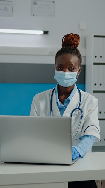 Portrait d'un médecin afro-américain assis au bureau avec masque facial dans un cabinet médical. Femme noire avec profession médicale utilisant un ordinateur portable moderne et portant un manteau blanc avec stéthoscope