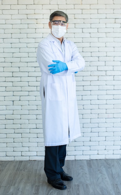 Portrait d'un médecin âgé asiatique de pleine longueur ou d'un chercheur portant une blouse de laboratoire, des lunettes claires et un masque facial debout et le bras croisé avec un fond de brique blanche.