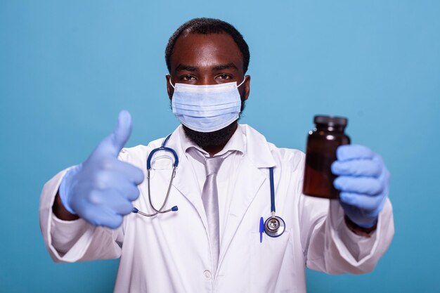 Portrait d'un médecin donnant le pouce en l'air tenant une bouteille de pilule portant un masque facial et des gants en latex. Médecin avec stéthoscope faisant un geste de la main approuvant les analgésiques sur ordonnance.