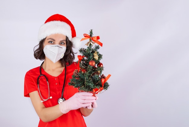 Portrait d'un médecin une femme dans un masque de protection médicale Bonnet de Noel et gants en nitrile rose tenant un arbre de Noël