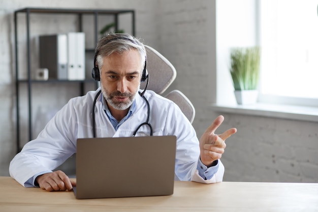 Photo portrait d'un médecin de sexe masculin aux cheveux gris dans son bureau utilisant un ordinateur portable pour une conversation vidéo avec un patient. consultation en ligne avec un médecin pour des diagnostics et des recommandations de traitement. concept de télésanté.
