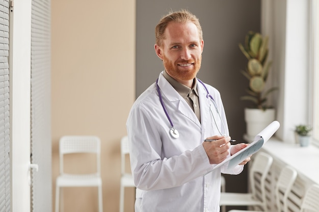 Portrait de médecin de sexe masculin barbu en blouse blanche tenant une carte médicale et souriant à l'avant debout dans le couloir de l'hôpital