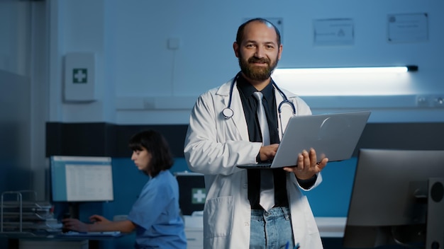 Portrait d'un médecin souriant portant un manteau médical et un stéthoscope tenant un ordinateur portable, tapant l'expertise en soins de santé. Personnel clinique travaillant pendant des heures au bureau de l'hôpital. Service de médecine