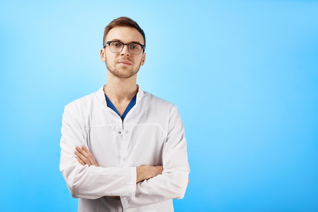 Portrait de médecin stagiaire en blouse médicale blanche et lunettes avec une expression de visage sérieux isolé sur fond de mur bleu