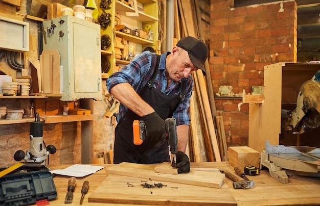 Portrait d'un menuisier senior avec instrument de forage travaillant dans l'atelier de menuiserie pour fabriquer un meuble