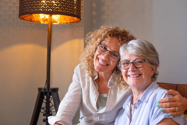 Portrait d'une mère âgée sereine et souriante et d'une fille blonde mature, étreignant assis sur un canapé à la maison