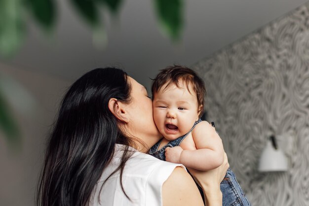 Portrait de mère aimante avec petite fille dans la chambre gros plan arrière-plan flou flou artistique Embrasser et embrasser le mécontentement bébé capturer une émotion heureuse Concept d'affection maternelle et de garde d'enfants