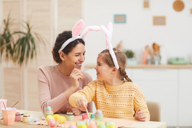 Portrait de mère aimante avec petite fille s'amusant tout en peignant des œufs de Pâques dans un intérieur de cuisine confortable, tous deux portant des oreilles de lapin, copiez l'espace